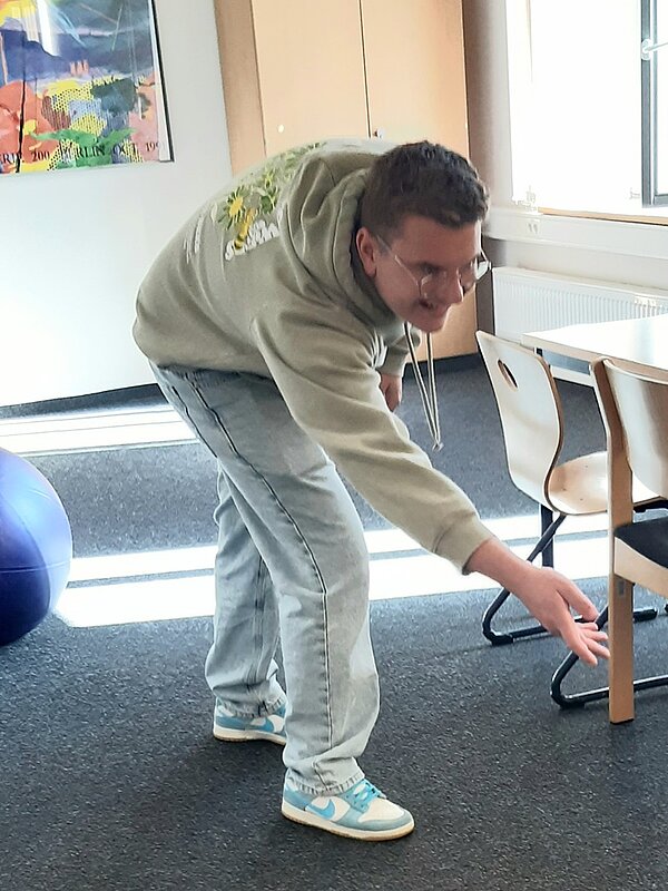 Einstimmung auf Ostern Ein männlicher Auszubildender in hellem Kapuzenpullover, mit kurzen braunen Haaren, hellen Nike Sneakers und einer Brille steht im Klassenraum und macht eine Bowlingbewegung, indem er in die Knie geht und die Hand nach vorn ausstreckt.
