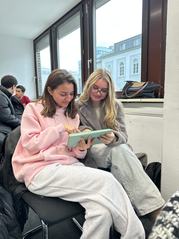 Zwei junge Frauen sitzen nebeneinander auf schwarzen Stühlen in einem hellen Raum mit großen Fenstern, durch die ein historisches Gebäude zu sehen ist. Die Frau links trägt einen hellrosa Pullover und eine hellgraue Hose, die Frau rechts einen grauen Pullover und eine hellgraue Hose. Beide konzentrieren sich auf ein hellgrünes Tablet, das die linke Frau hält, während die rechte Frau mit der rechten Hand darauf zeigt. Im Hintergrund sind weitere Personen zu erkennen, die ebenfalls auf Stühlen sitzen. Auf der Fensterbank liegt eine schwarze Tasche mit braunen Griffen. Die Szene vermittelt eine Lern- oder Workshop-Atmosphäre, passend zum Thema "Management Erleben" im Rahmen eines Workshops der ISM an der Euro Akademie Wiesbaden.