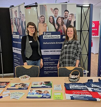Zwei weibliche Mitarbeitende der Euro Akademie stehen lächelnd an ihrem Messestand.