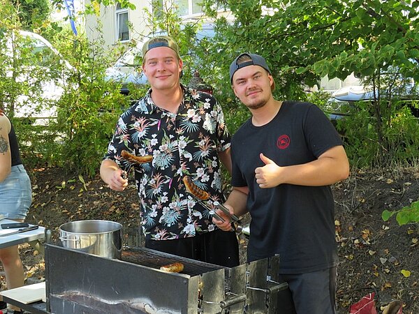 Zwei gute gelaunte, männliche Auszubildende grillen Würstchen für die Besucher*innen.