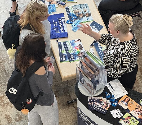 Eine Mitarbeiterin der Euro Akademie mit hellen Haaren und auffälligem Haarclip sitzt hinter dem Messestand und spricht mit zwei jungen Menschen, die vor dem Stand stehen. Der Stand ist mit Broschüren, Flyern und kleinen blauen Werbeartikeln bestückt. Auf dem Tisch liegen weitere Informationsmaterialien und ein blaues Stofftäschchen mit weißem Schriftzug. Die Szene spielt in einem Innenraum mit hellem Boden.