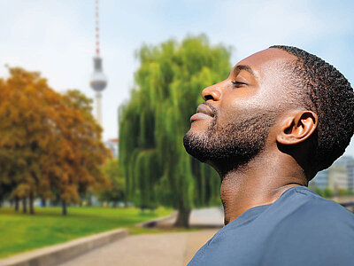 Ein Mann steht entspannt in einem Park und genießt die Natur. Er trägt ein blaues T-Shirt und hat die Augen geschlossen, während er den Kopf leicht nach oben neigt, als würde er die Sonne oder die frische Luft spüren. Im Hintergrund sind grüne Bäume, eine Wiese und der Fernsehturm in Berlin zu sehen. Die Szene vermittelt eine friedliche und erholsame Atmosphäre. Der Mann wirkt zufrieden und entspannt.