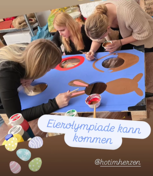 Mehrere Personen bemalen gemeinsam ein großes Spielbrett mit Osterhasen- und Eiermotiven für die Eierolympiade.