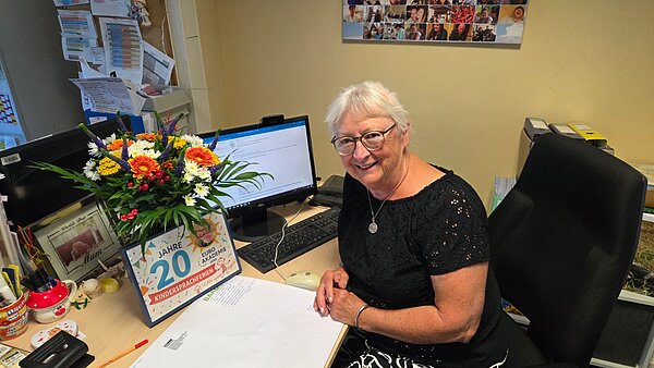 Sprachferien der Euro Akademie Jena und Erfurt: eine 20-jährige Erfolgsgeschichte Eine Frau sitzt am Schreibtisch und blickt freundlich in die Kamera. Auf dem Schreibtisch steht ein Laptop, ein Blumenstrauß und ein Jubiläumsgeschenk in Form eines Bilderrahmens