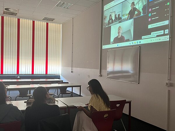 Studierende sitzen in einem Unterrichtsraum und verfolgen eine zugeschaltete Online-Präsentation, die an die Wand projiziert wird.