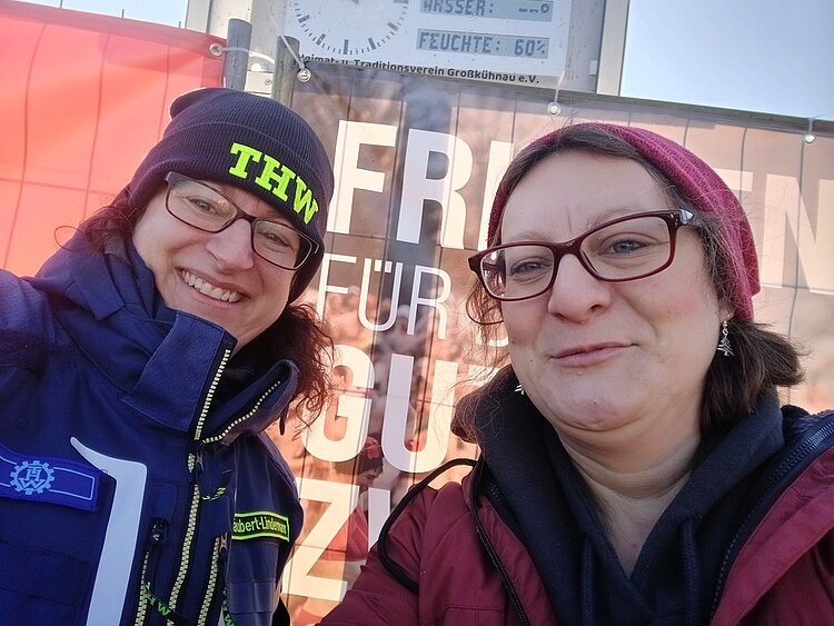 Frieren für den guten Zweck: unterstützt durch die Klasse EZ 23 Die Lehrkraft, nun vollständig bekleidet mit Jacke und Mütze, grinst zusammen mit einer Freiwilligen des THW für ei Selfie in die Kamera.