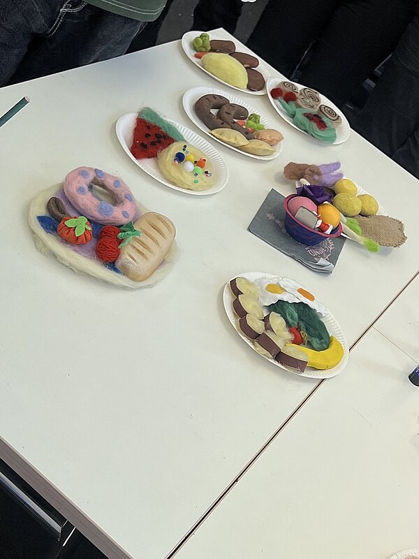 Auf einem weißen Tisch liegen mehrere Pappteller, die mit farbenfrohen Filzobjekten belegt sind. Die Filzobjekte stellen verschiedene Speisen dar, darunter Brot, Obst, Gemüse, ein Donut, Kekse und eine Melonenscheibe. Die Filzspeisen sind detailreich gestaltet und in kräftigen Farben gehalten. Im Hintergrund stehen mehrere Personen, deren Beine und Hosen sichtbar sind.