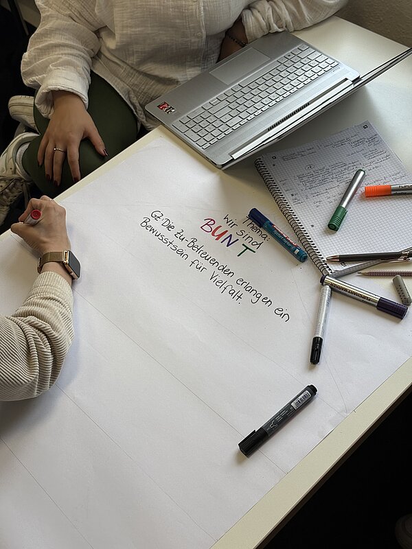 Foto von einem Tisch, auf dem ein großes weißes Papier und mehrere Stifte liegen; jemand schreibt gerade darauf; auf dem Plakat steht "wir sind bunt"