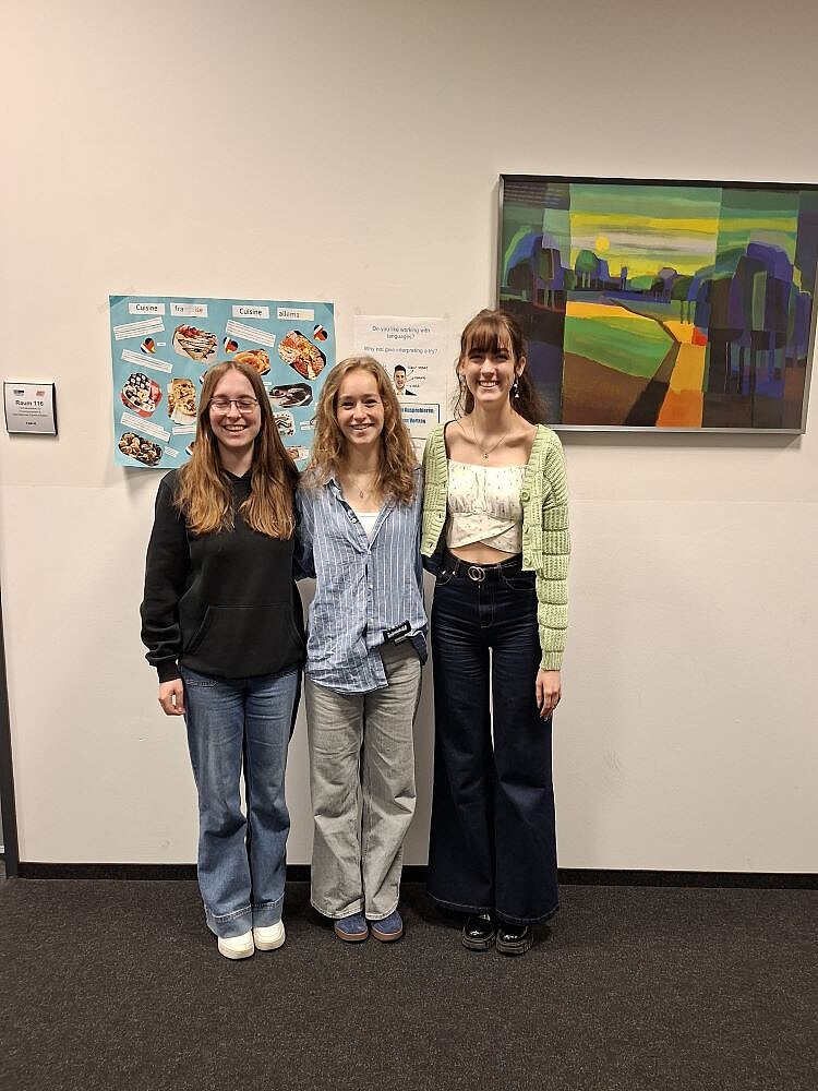 Drei lächelnde junge Frauen stehen nebeneinander in einem Flur. Die Person links trägt ein schwarzes Sweatshirt und Jeans, die Person in der Mitte ein hellblaues Hemd und graue Hosen, und die Person rechts ein hellgrünes Cardigan-Top, eine dunkle Hose. Im Hintergrund hängen ein Poster mit Speisenabbildungen und ein buntes Landschaftsgemälde an der Wand.