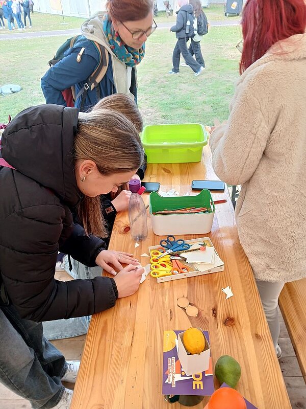 Eine Person bastelt konzentriert an einem Tisch im Freien. Auf dem Tisch liegen Schere, Kleber, Papier und andere Bastelutensilien. Im Hintergrund sind weitere Personen zu sehen, die ebenfalls an Aktivitäten teilnehmen.
