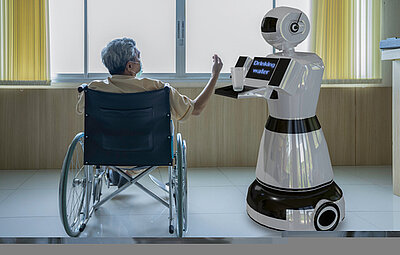 Ein älterer Mensch sitzt im Rollstuhl und interagiert mit einem Pflegeassistenzroboter. Der Roboter hält ein Tablett mit einer Tasse und zeigt auf einem Display den Text "Drinking water" an. Die Szene spielt sich in einem hellen Raum mit großen Fenstern und gelben Vorhängen ab.