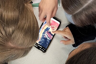 Drei Jugendliche beugen sich über einen Tisch und betrachten gemeinsam ein Smartphone, auf dessen Bildschirm ein farbiges Foto einer Person mit Krone und auffälligem Kostüm zu sehen ist. Zwei Hände zeigen mit ausgestrecktem Finger auf Details des Bildes. Die Szene vermittelt konzentrierte Zusammenarbeit und aktives Diskutieren, passend zum Projekt „Werde zum Faktenchecker“. Die Aufnahme betont den interaktiven Charakter und die digitale Recherche im Team.