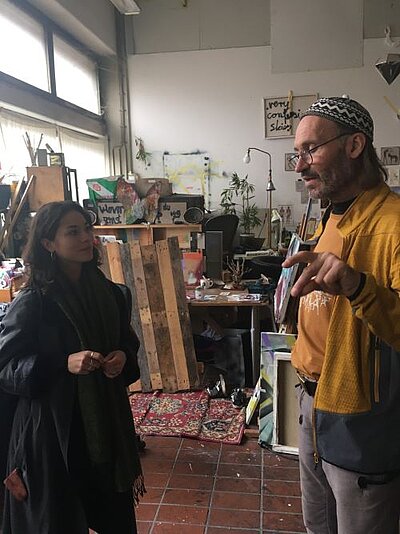 Eine junge Frau und ein Mann unterhalten sich in einem Atelier. Der Mann trägt eine gemusterte Mütze, eine Brille und eine gelbe Jacke, während er gestikulierend spricht. Die Frau steht ihm gegenüber, hört aufmerksam zu und hält etwas in ihren Händen. Im Hintergrund sind kreative Werke, Holzpaletten, Pflanzen und verschiedene Kunstmaterialien zu sehen, die eine inspirierende Arbeitsatmosphäre schaffen.