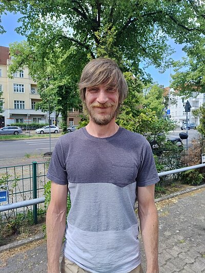 Portraitbild von Thomas Bernardy, Alumnus der Euro Akademie Berlin. Er steht im Freien. Im Hintergrund sind Bäume, ein Häuser und Autos zu erkennen.