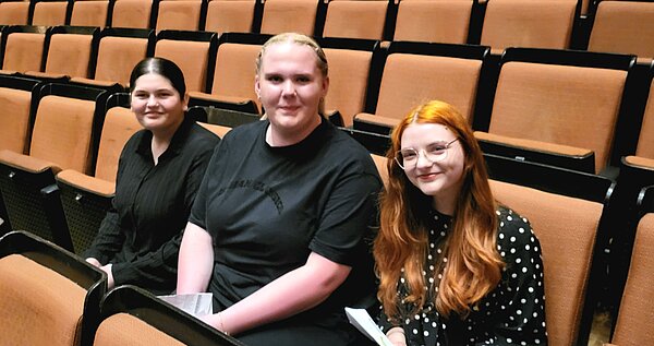All That Jazz! Englischunterricht im Opernhaus Hannover Drei Personen sitzen nebeneinander in einem Theatersaal mit orangefarbenen Stühlen. Alle lächeln in die Kamera.