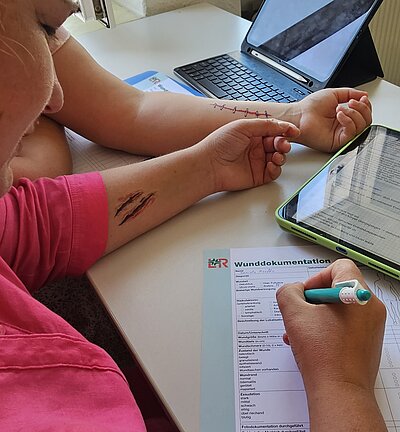 Zwei Personen sitzen an einem Tisch, eine dokumentiert auf einem Formular „Wunddokumentation“. Auf den Armen der anderen Person sind realistisch aussehende Wunden und eine Naht als Übungsdarstellung zu sehen.