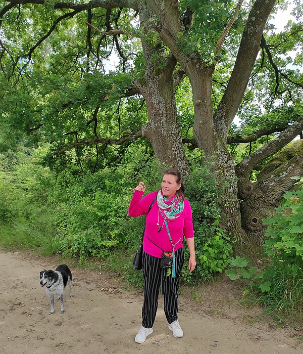 Walk & Talk Eine Frau in einem leuchtend pinken Pullover und einer schwarzen Hose steht vor einer mächtigen alten Eiche, daneben ein halbhoher schwarz-weiß melierter Hund. Die Frau scheint gerade etwas zu erklären, da sie die rechte Hand während des Sprechens erhebt.