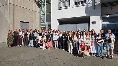 Eine große Gruppe von jungen Erwachsenen posiert lächelnd vor dem Eingang eines Gebäudes mit der Aufschrift „Thomasstrasse 83“. Die Stimmung wirkt fröhlich und entspannt.