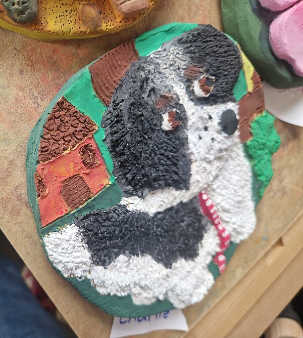 Ein rundes Tonrelief mit einem Hund in Schwarz-Weiß, der auf einer grünen Wiese vor einem stilisierten Haus und einem Baum steht. Das Relief liegt auf einer Holzplatte. 