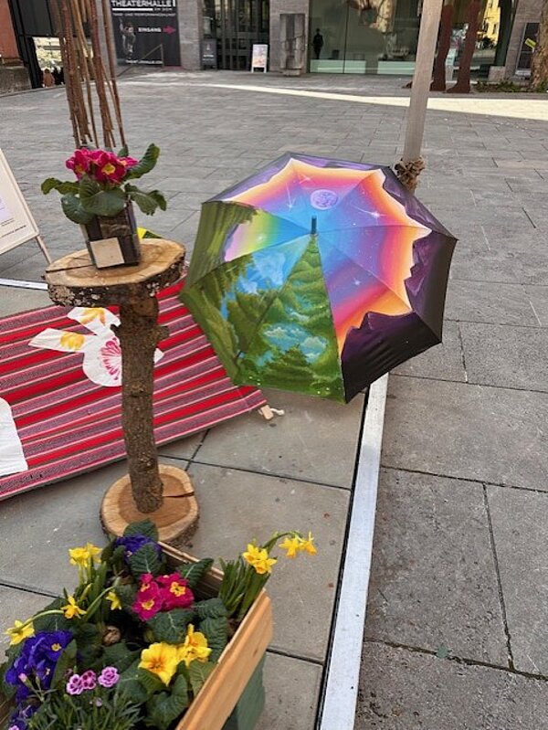 Auf einem Platz vor dem Dom ist Kunst ausgestellt: darunter ein umgedrehter Regenschirm mit Blumen darin; ein bunt bemalter Regenschirm und eine Hängematte mit dem Wort "Ruhe".