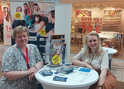 Zwei Mitarbeiterinnen am Messestand der Euro Akademie Dessau posieren gut gelaunt an einem Tisch, auf dem Infoflyer liegen, für ein Bild.