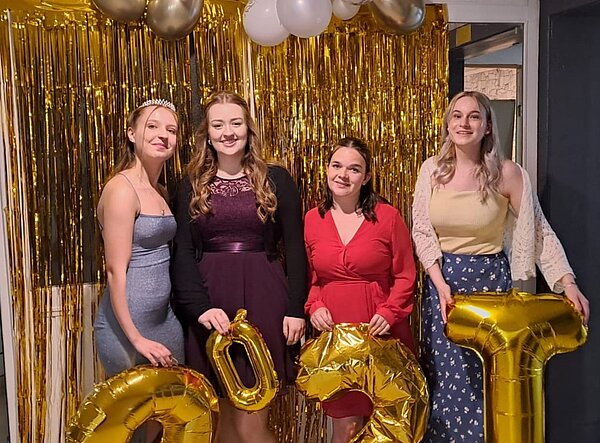 Feierliche Übergabe der Zeugnisse Vier junge Mädchen in Kleidern bzw. einem langen Rock stehen vor einem Vorhang aus goldenem Lametta. Von der Decke hängen weiße und goldene Luftballons. Die Mädchen halten goldene Luftballons mit Zahlen in unterschiedlicher Größe in der Hand. Das Mädchen links hat eine Krone im Haar.