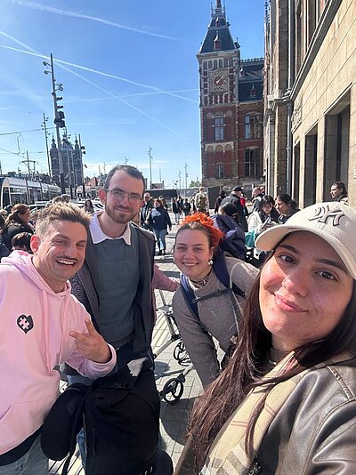 Eine Gruppe von vier Personen posiert lächelnd für ein Foto auf einer belebten Straße in Amsterdam. Im Hintergrund sind historische Gebäude und eine Straßenbahn zu erkennen.