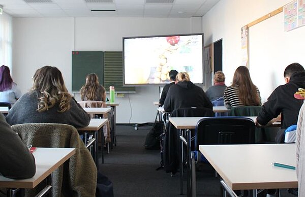 Theorie meets Praxis Ein Seminarraum mit einer Tafel, einem Monitor, Tischen und Stühlen, auf denen Teilnehmende von hinten zu sehen sind.