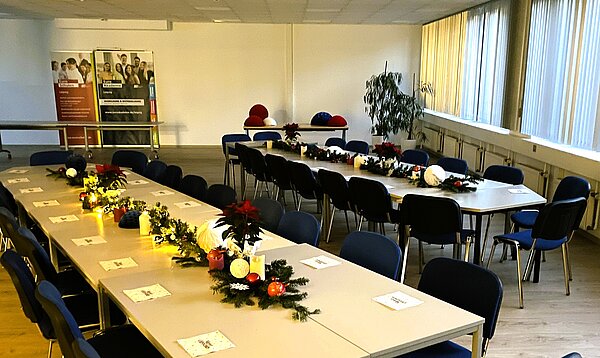 Großes Auditorium mit langen, zu U-Form gestellten Tischen, blauen Stühlen und weihnachtlicher Tischdekoration aus Lichterketten, Tannengrün und roten Sternen.