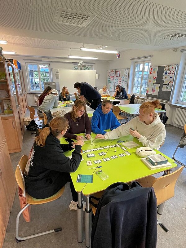 Vier Personen um einen grünen Tisch in einem hellen Klassenzimmer, gemeinsam spielen sie ein Brettspiel mit kleinen bunten Elementen. Im Hintergrund sind weitere Gruppen an Tischen. Zudem sind Regale, bunte Bilder an der Wand und ein Whiteboard zu sehen.