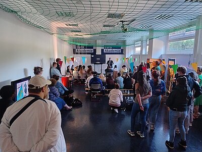 Stehende und sitzende Besucherinnen und Besucher hören der Rede des stellvertretenden Schulleiters in den Räumlichkeiten der Euro Akademie zu.