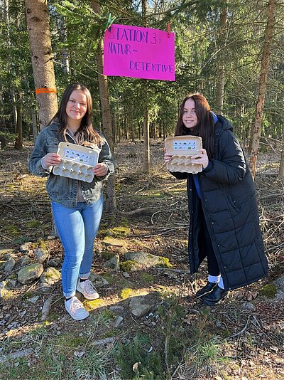 Zwei junge Frauen stehen in einem lichten Waldstück unter einem pinken Schild mit der Aufschrift „Station 3: Naturdetektive“. Sie halten Eierkartons mit kleinen Bildern, die als Sammelhilfe für Naturmaterialien dienen. Beide lächeln in die Kamera.