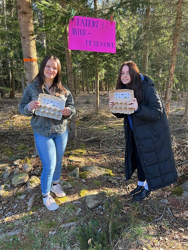 Zwei junge Frauen stehen in einem lichten Waldstück unter einem pinken Schild mit der Aufschrift „Station 3: Naturdetektive“. Sie halten Eierkartons mit kleinen Bildern, die als Sammelhilfe für Naturmaterialien dienen. Beide lächeln in die Kamera.