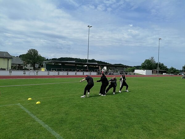Vier Auszubildende der Euro Akademie Zwickau ziehen gemeinsam an einem Seil auf einer Rasenfläche. Sie tragen dunkle Kleidung und stehen hintereinander in einer Linie. Im Hintergrund sind eine Tribüne, einige Gebäude und Bäume zu sehen. Der Himmel ist teilweise bewölkt. Auf dem Rasen liegen zwei gelbe Markierungshütchen.