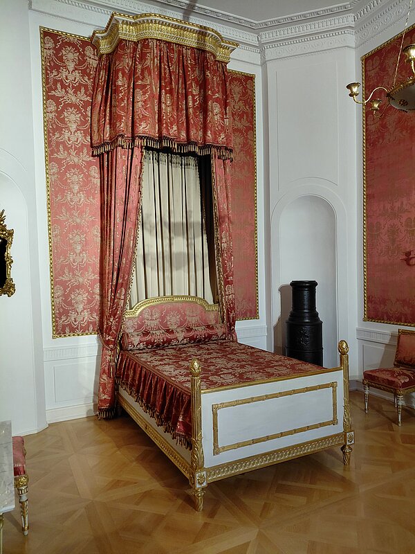 Eintauchen in die Geschichte Aschaffenburgs Ein reich verziertes Bett mit rotem Brokatstoff und goldenen Elementen steht in einem eleganten Schlosszimmer mit weißen Wänden, rotem Wandbehang und Parkettboden.