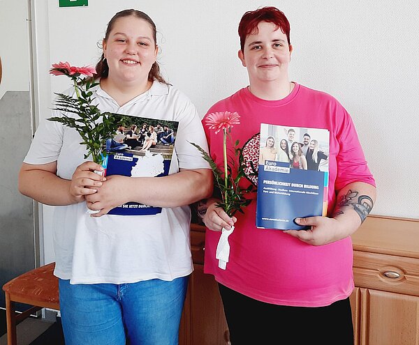 Pflegehelfer*innen in Bitterfeld-Wolfen verabschiedet Zwei Schülerinnen freuen sich über ihre erfolgreich abgeschlossene Ausbildung. Sie halten mit einem breiten Grinsen ihre Zeugnismappen in die Kamera.
