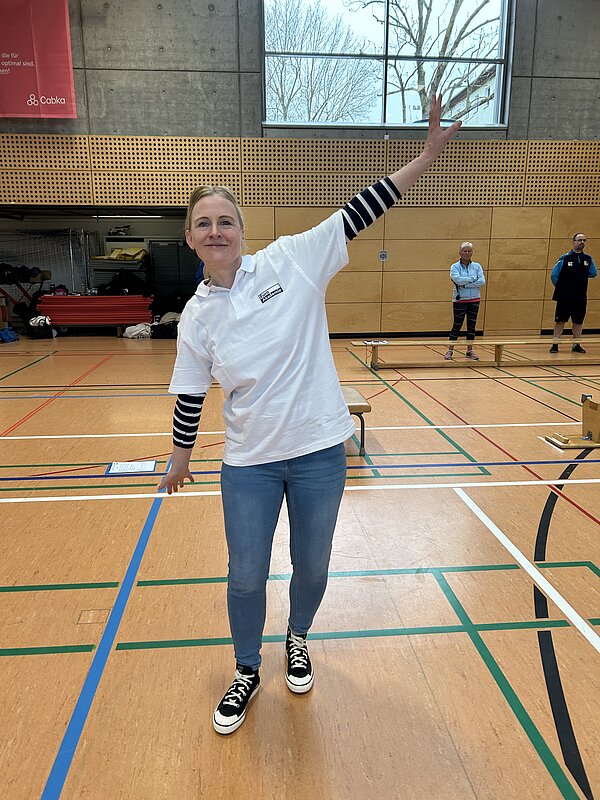 Auszubildende der Euro Akademie Pößneck unterstützen erneut den „Pokal des Bürgermeisters“ In einer Turnhalle steht eine blonde Frau in einem weißen T-Shirt mit Logo der Euro Akademie, die ihre Arme wie ein Flieger ausbreitet. Sie trägt eine blaue Jeans und schwarze Sneaker. Im Hintergrund stehen weitere Personen hinter einer Holzbank, daneben ist hinter einer geöffneten Garagentür ein Materialraum erkennbar.