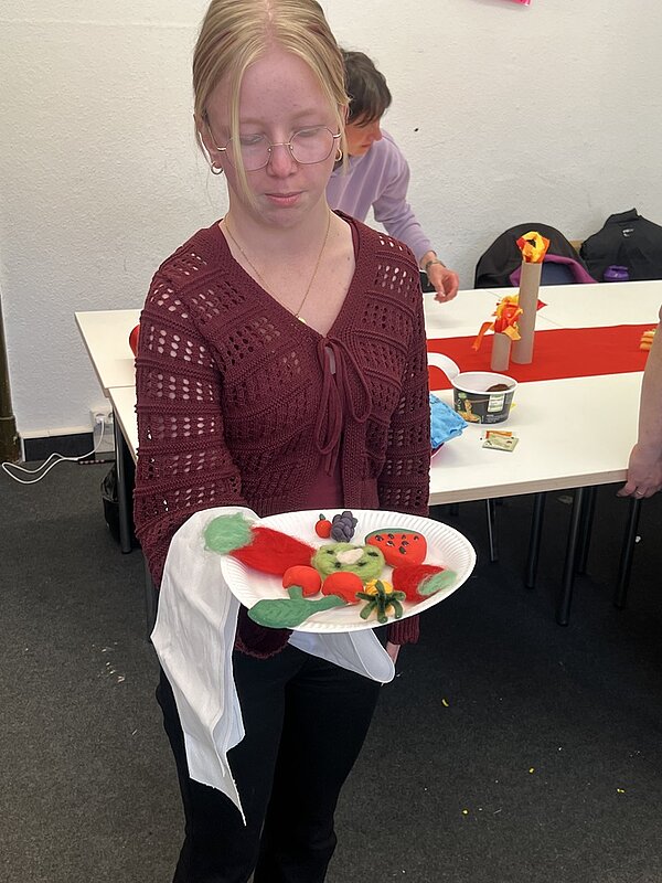 Eine Auszubildende zeigt stolz ihre kreativen Bastelarbeiten, diverses Obst aus Filz, in die Kamera.