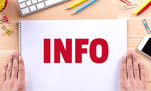 Die Aufschrift "Info" in großen roten Lettern steht auf einem Zeichenblock geschrieben, der auf einem Holztisch liegt und neben dem rechts und links die Hände einer Person flach aufliegen. Um den Block verteilt liegen Stifte, Büroklammern und ein Handy und es ist die Ecke einer Tastatur zu sehen.