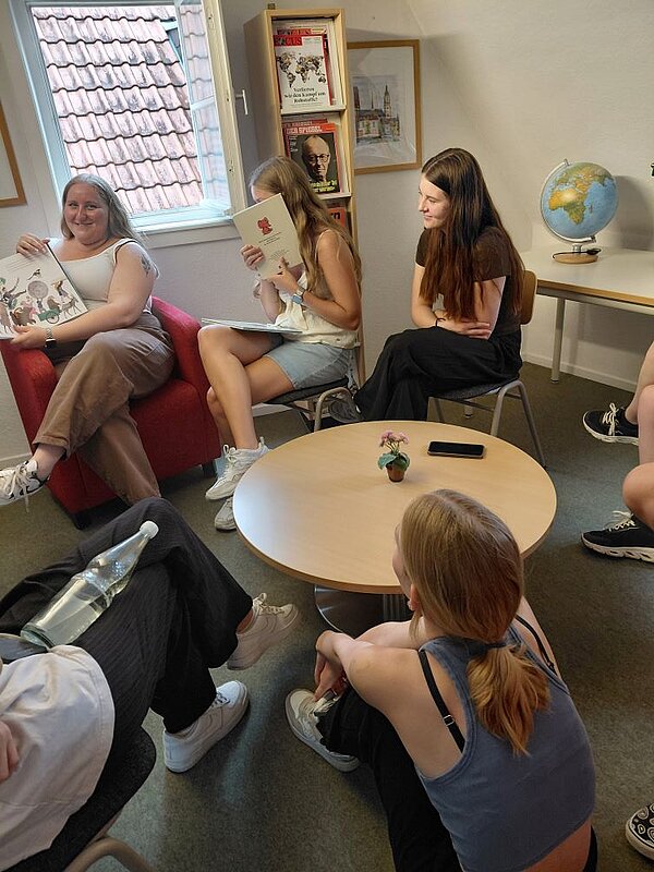 Eine Gruppe von jungen Erwachsenen sitzt um einen runden Tisch in einem Raum mit Büchern und Spielzeug. Eine Person liest ein Buch, während die anderen sich unterhalten. Im Hintergrund sind Regale mit Büchern und ein Globus zu erkennen.