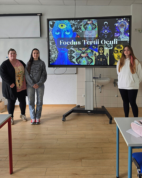 Drei Personen posieren vor einem Bildschirm, auf dem eine Präsentation mit dem Titel „Foedus Tertii Oculi“ zu sehen ist. Die Folie enthält mystische Symbole und Augen.