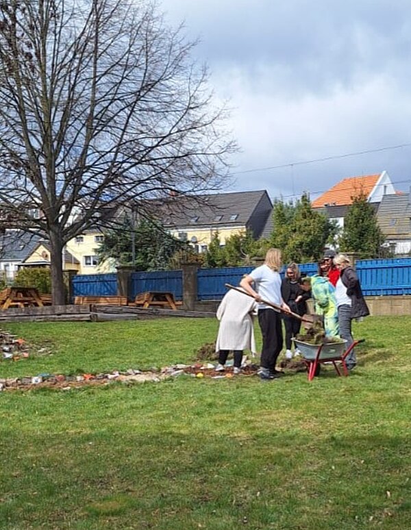 Mehrere Auszubildende stehen auf einer Rasenfläche und arbeiten gemeinsam an einem Beet, das mit Steinen eingefasst ist. Eine Schubkarre steht neben ihnen. Im Hintergrund sind ein großer, kahler Baum, eine Mauer, Holzbänke und Wohnhäuser mit Satteldächern zu sehen. Der Himmel ist bewölkt.