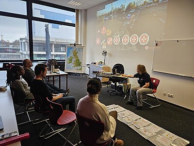 Erste-Hilfe-Schulung im Seminarraum: Trainerin zeigt eine Folie mit Verkehrsunfall-Symbolen, fünf Teilnehmende sitzen zugewandt; auf dem Boden liegen Ablaufkarten, neben der Leinwand steht ein Flipchart.