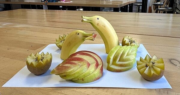 Bananen sind als Delphine dargestellt, mit einem kunstvoll zugeschnittenen Apfel und einer Kiwi garniert. 