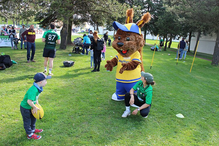 Ein Kind mit Ball steht auf einer Wiese vor einem Maskottchen und einer Betreuungsperson; im Hintergrund finden weitere Bewegungsangebote statt. Das Bild hat uns der Sportbund zur Verfügung gestellt.