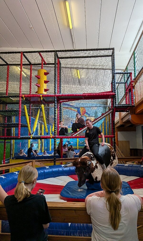 Eine junge Frau sitzt auf einem mechanischen Rodeo-Bullen mit braun-weißem Fell. Das Luftkissen darunter hat rot-weiße Streifen und blaue Flächen mit weißen Sternen. Im Vordergrund stehen Zuschauerinnen. Im Hintergrund sind Klettergerüste und Netze zu sehen, in denen weitere junge Menschen sitzen.