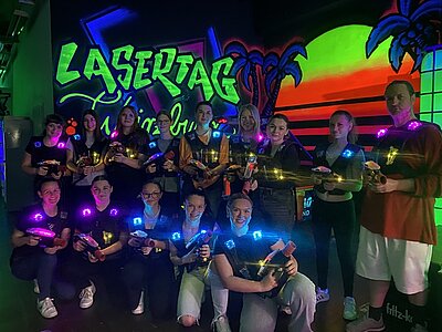 Junge Menschen stehen in zwei Reihe, teilweise gebückt, in einem dunkeln Raum. Sie werden von kleinen bunten Lichtern an ihren Oberteilen beleuchtet und halten Lasertag-Pistolen in ihren Händen. Im Hintergrund ist der Schriftzug "Lasertag" erkennbar, sowie ein Sonnenunterang mit Palmen als Wandbild.