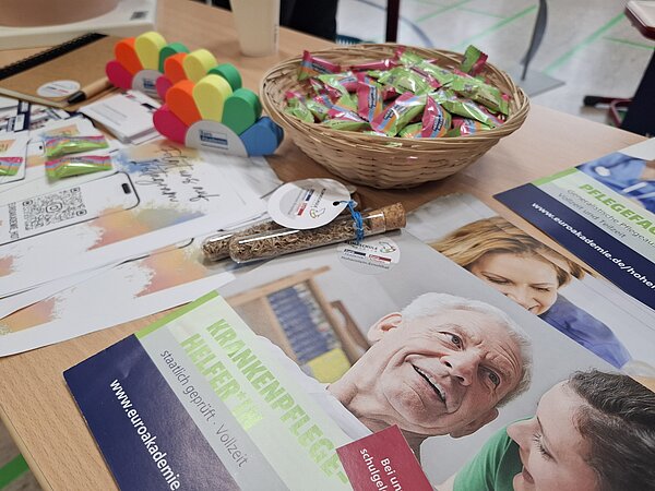 Informationsmaterialien zur Pflegeausbildung liegen mit kleinen Give-aways und Süßigkeiten auf einem Messetisch.