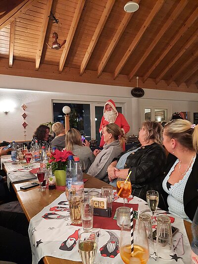 Weihnachtsfeier in gemütlicher Gaststube: Nikolaus spricht mit Gästen an einem langen, festlich gedeckten Tisch mit vielen Gläsern und Deko.
