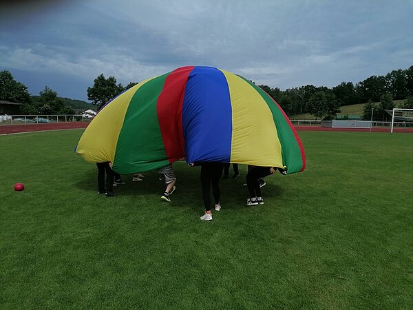 Auf dem große Rasensportplatz halten mehrere Kinder ein buntes Schwungtuch, das in die Höhe geworfen wurde und eine halbkugelförmige Kuppel bildet. Die Farben des Tuchs sind gelb, grün, blau und rot. Unter dem Tuch sind nur die Beine der Kinder sichtbar. Im Hintergrund befinden sich eine Laufbahn, ein Fußballtor, Bäume und einige Gebäude.